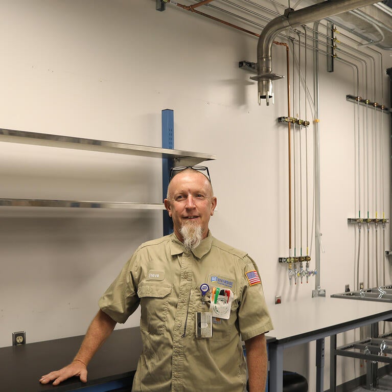 A technician standing inside a clean laboratory with overhead piping and wall-mounted valves
