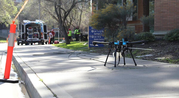 A black drone sits on the sidewalk next to a brick building with a van in the background