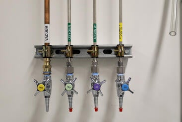 A wall-mounted laboratory gas manifold distributing vacuum, oxygen, argon, and nitrogen through four labeled, color-coated lines